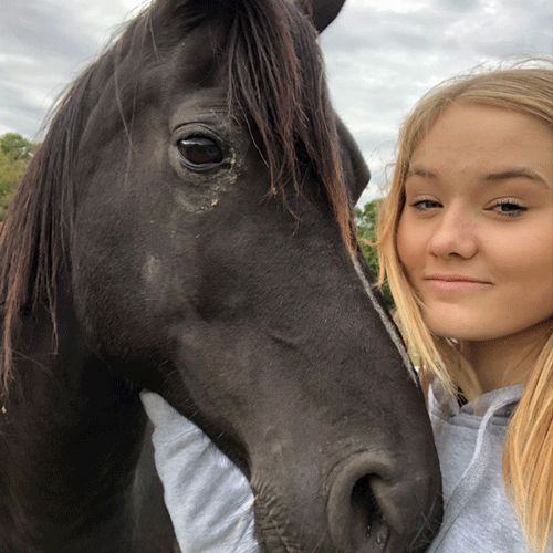 Carly, a Certified Dog Handler at The Chase, poses with a black horse on a cloudy day.