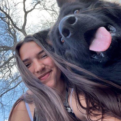 Kailee, a Certified Dog Handler at The Chase, takes a selfie with a black dog whose pink tongue is out.